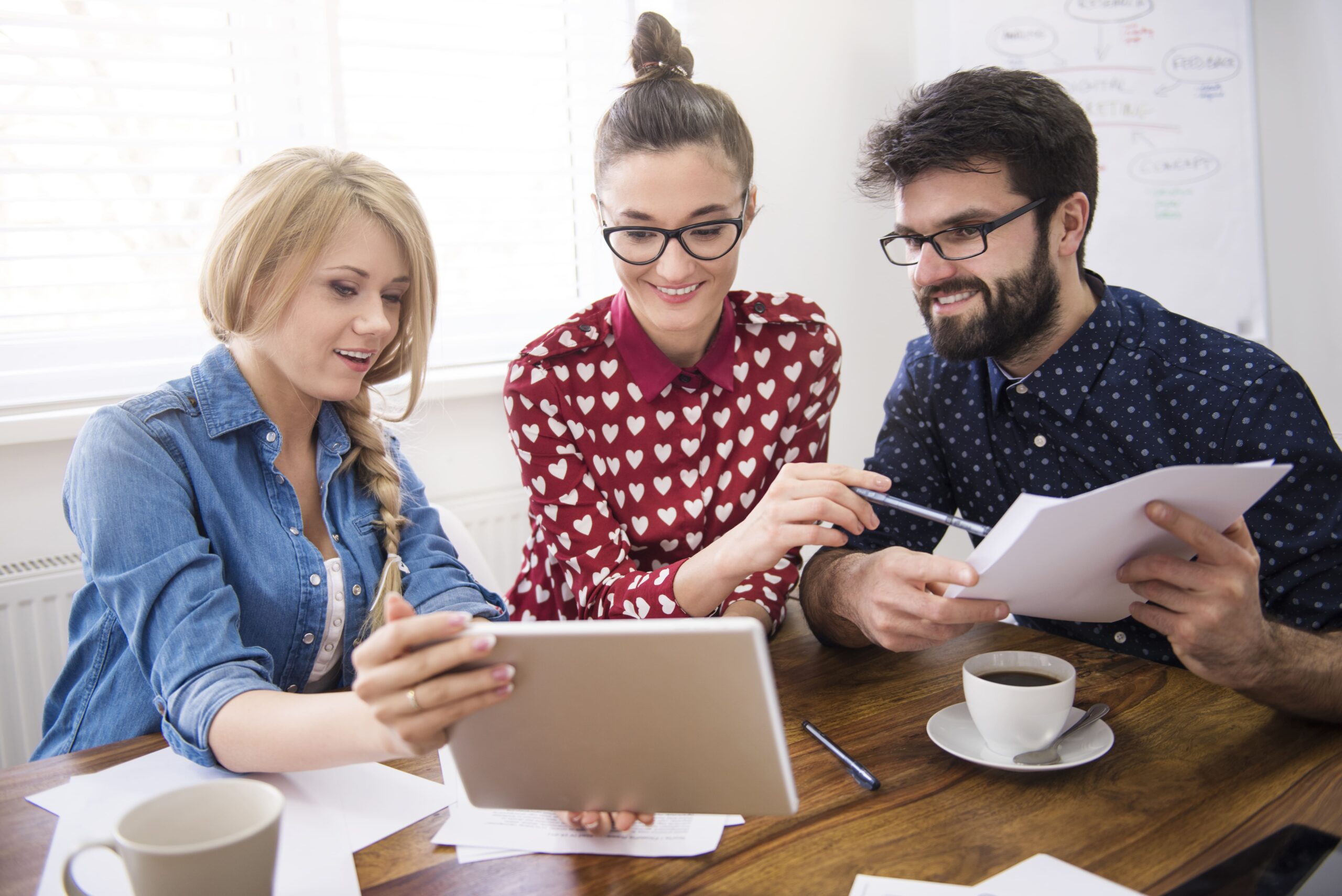 Tres personas reunidas en una mesa de trabajo, observando y señalando una tablet mientras comparten ideas y papeles. Hay tazas de café en la mesa.