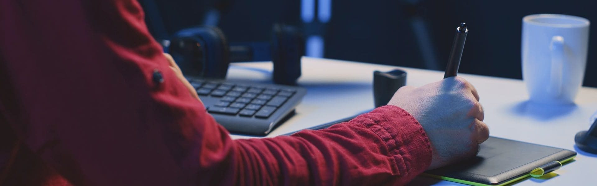 Banner de infografía, La imagen muestra la mano de un hombre utilizando un Apple Pencil sobre una tableta gráfica que está sobre un escritorio blanco. En el escritorio también se encuentran un pocillo blanco, un teclado negro de computadora y unos auriculares negros.
