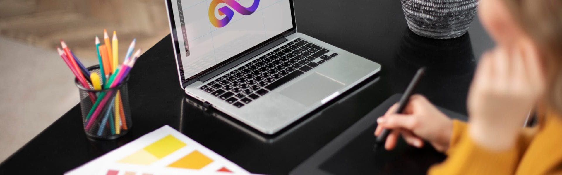 Banner de logotipos y branding, La imagen muestra un escritorio negro donde una mujer utiliza un Apple Pencil sobre una tableta gráfica. En el escritorio se encuentran varios elementos: una hoja con muestras de colores, un recipiente que contiene lápices de colores, un portátil abierto con el símbolo del infinito en la pantalla, y la base de un jarrón visible parcialmente.